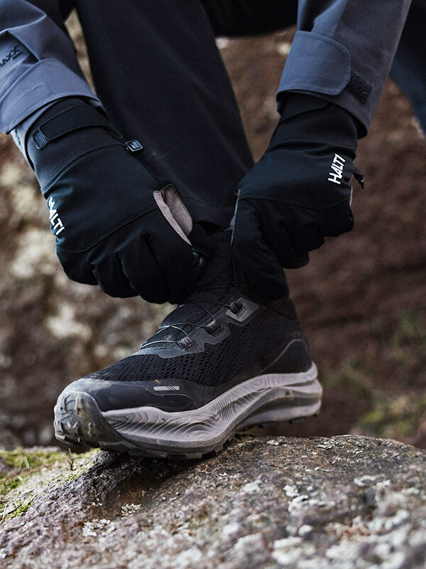 A person wearing black gloves tightens the laces on a black athletic shoe while standing on a rough, rocky surface outdoors.