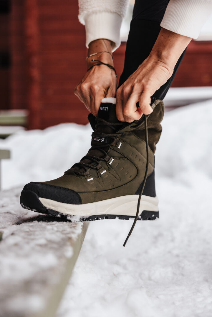 A person wearing a cream sweater is tying the laces on an olive green winter boot while standing on a snow-covered surface.