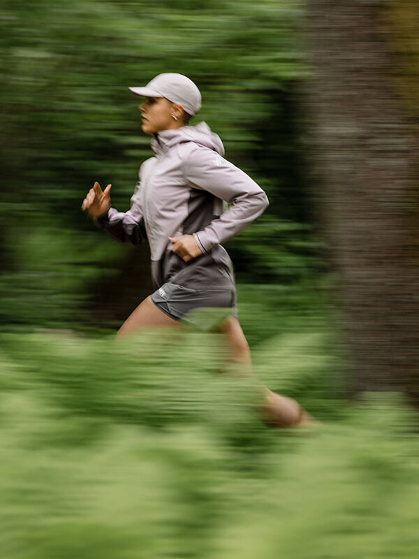A person wearing a light-colored cap and jacket runs outdoors through a green, blurred natural environment. The image captures motion, showing the runner in mid-stride.