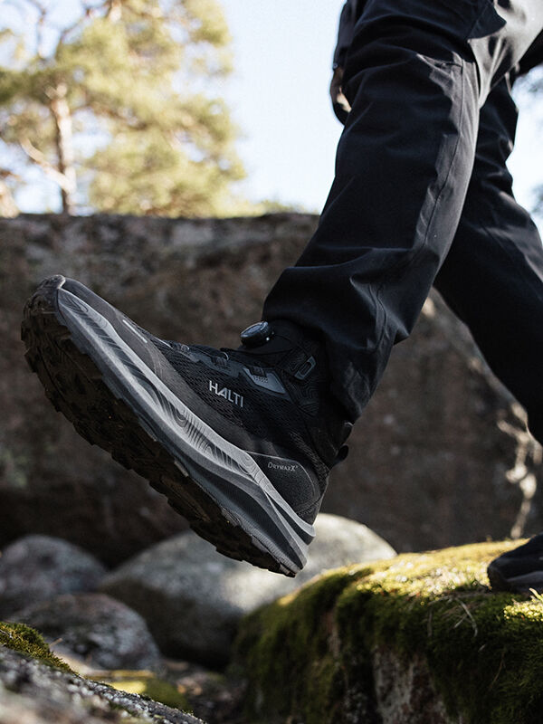 A person wearing black outdoor pants and black Halti hiking shoes is stepping on moss-covered rocks, with trees and more rocks in the background.