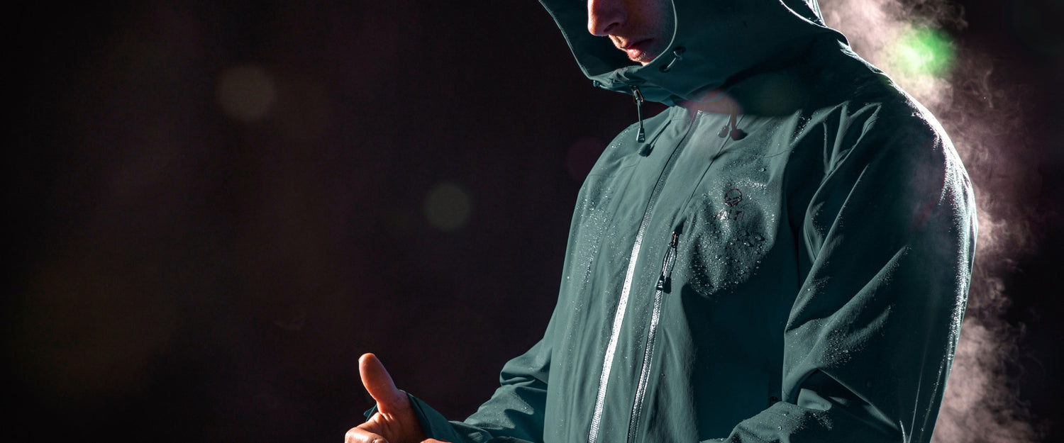 A person wearing a dark green waterproof jacket with the hood up is shown against a dark background, with visible water droplets on the jacket and mist in the air.