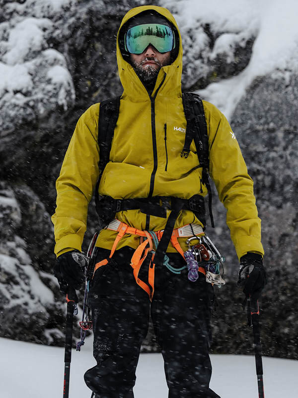 A person wearing a yellow jacket, black pants, and ski goggles stands in a snowy mountain environment, equipped with climbing gear and trekking poles. Snow is falling lightly.