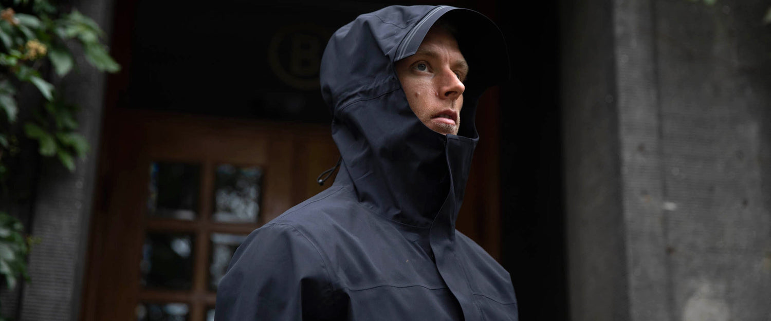 A person wearing a dark waterproof hooded jacket stands outdoors in front of a wooden door and concrete wall, looking slightly to the left.