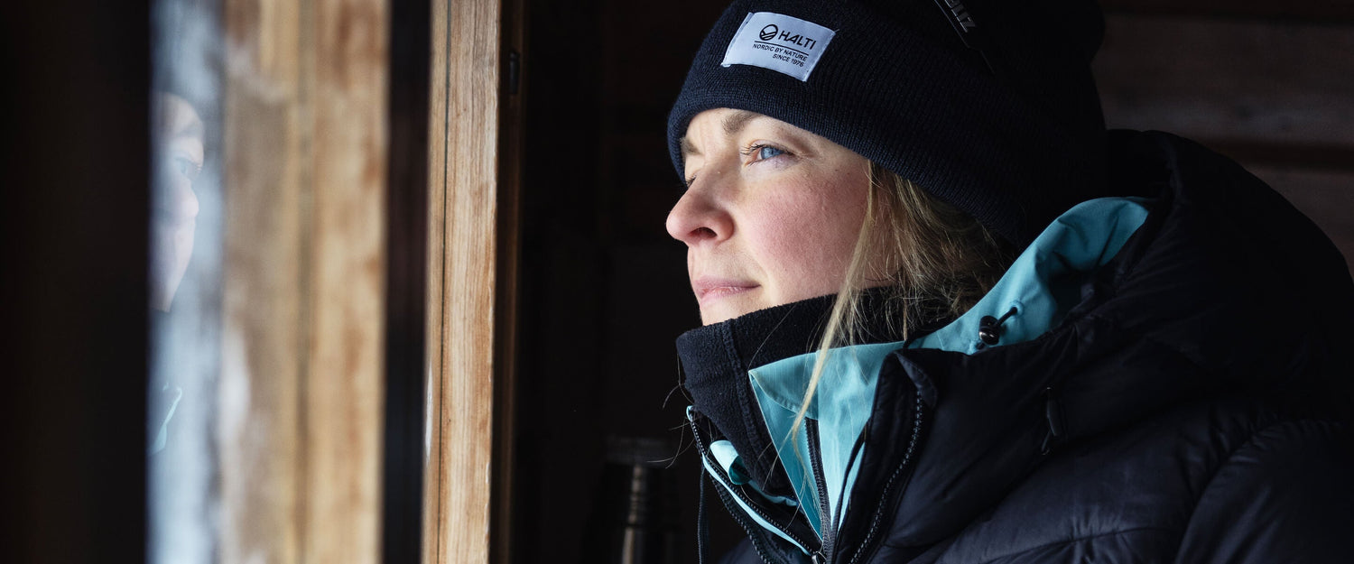 A woman wearing a black winter jacket and a black knit hat looks thoughtfully out of a window, with natural light illuminating her face.