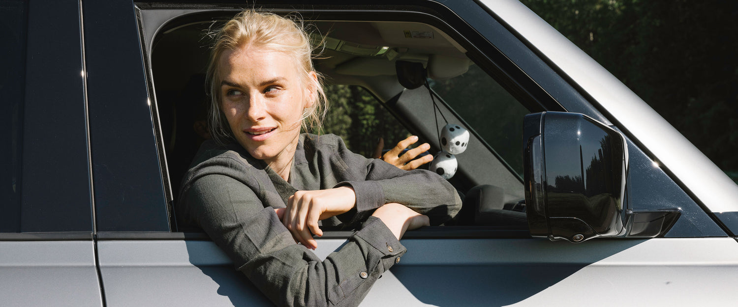 A woman with blonde hair sits in the driver’s seat of a silver SUV, resting her arms on the open window. A small panda toy hangs from the rearview mirror. Trees are visible in the background.