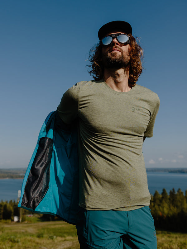 A man with long hair wearing sunglasses, a cap, and a light green t-shirt stands outdoors by a lake, pulling on a blue jacket with a forested landscape in the background.