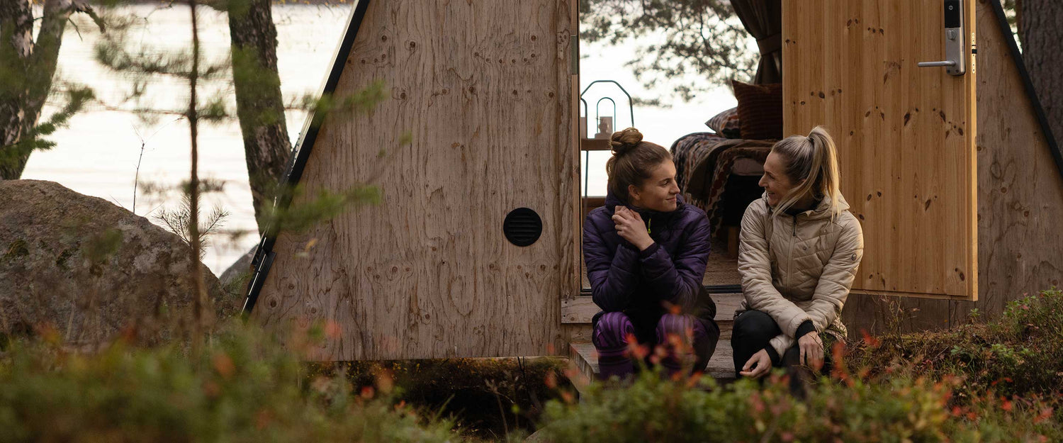 Two people sit on the wooden steps of a small A-frame cabin in a forested area, facing each other and talking. The open door reveals part of the cabin interior.