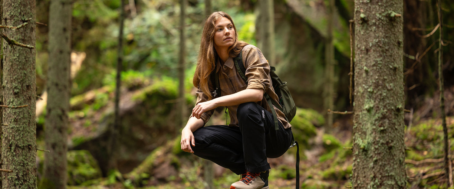 A person with long hair, wearing a brown shirt, black pants, and hiking boots, squats in a forest with trees and green moss in the background, carrying a green backpack.