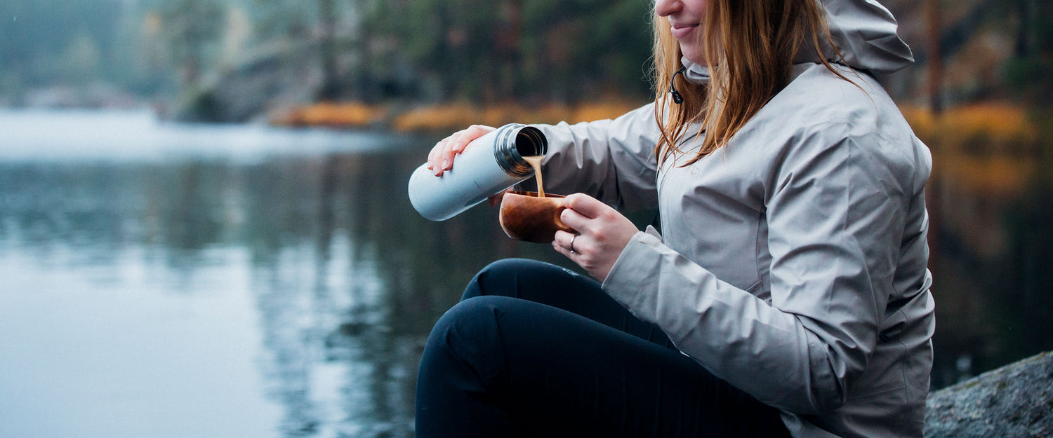 A person in a light gray jacket sits by a lake, pouring a drink from a thermos into a cup, with blurred trees and water in the background.