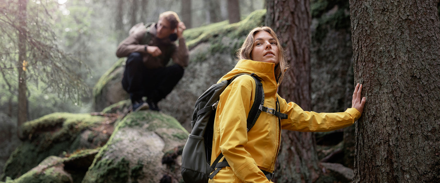 A woman in a yellow rain jacket and backpack touches a tree in a forest, while a man in the background squats on a mossy rock, surrounded by trees.