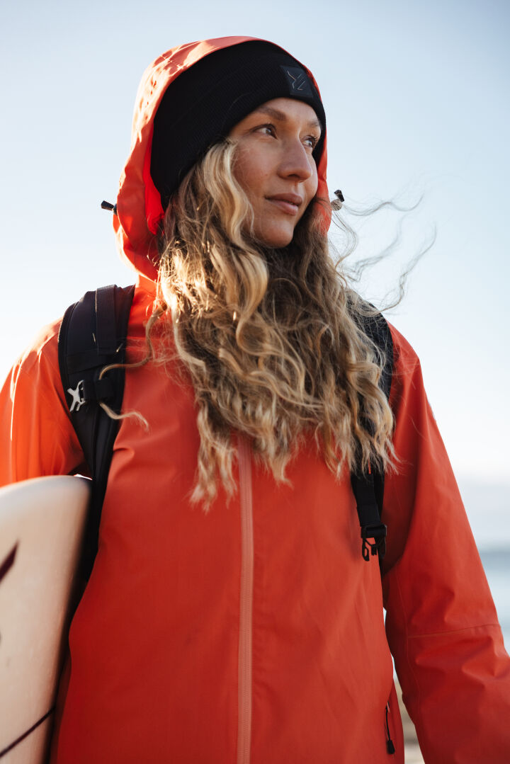 A person with long wavy hair wearing an orange jacket and black beanie stands outdoors, holding a surfboard and looking to the side. The sky in the background is clear.