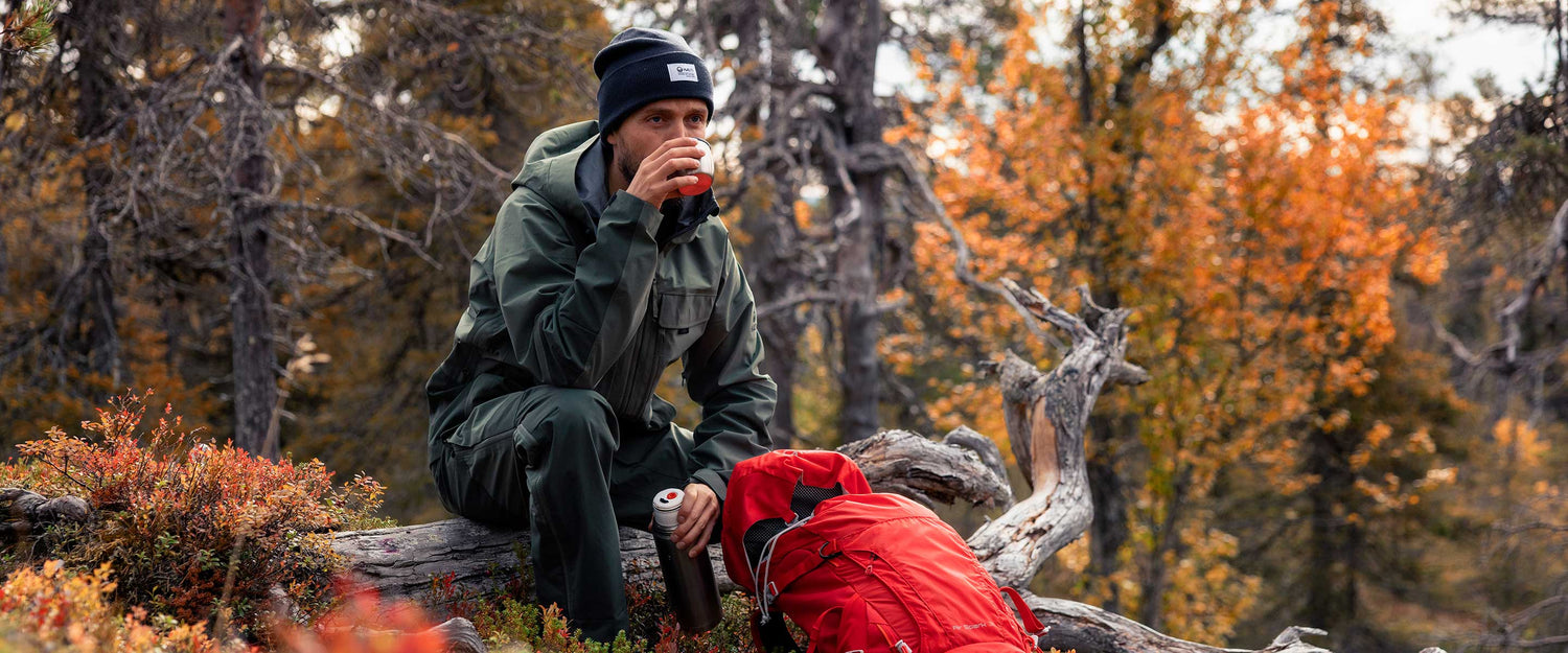 A person in green outdoor clothing and a dark beanie sits on a fallen tree, drinking from a cup. A red backpack and a thermos are nearby. Autumn trees fill the background.
