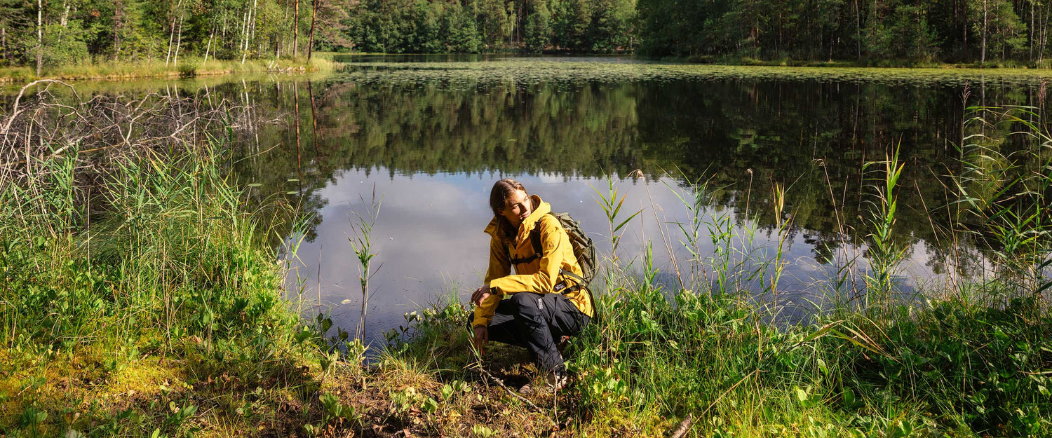 A person wearing a yellow jacket and backpack kneels by the edge of a calm forest lake, surrounded by green grass, trees, and reflections in the water under clear daylight.
