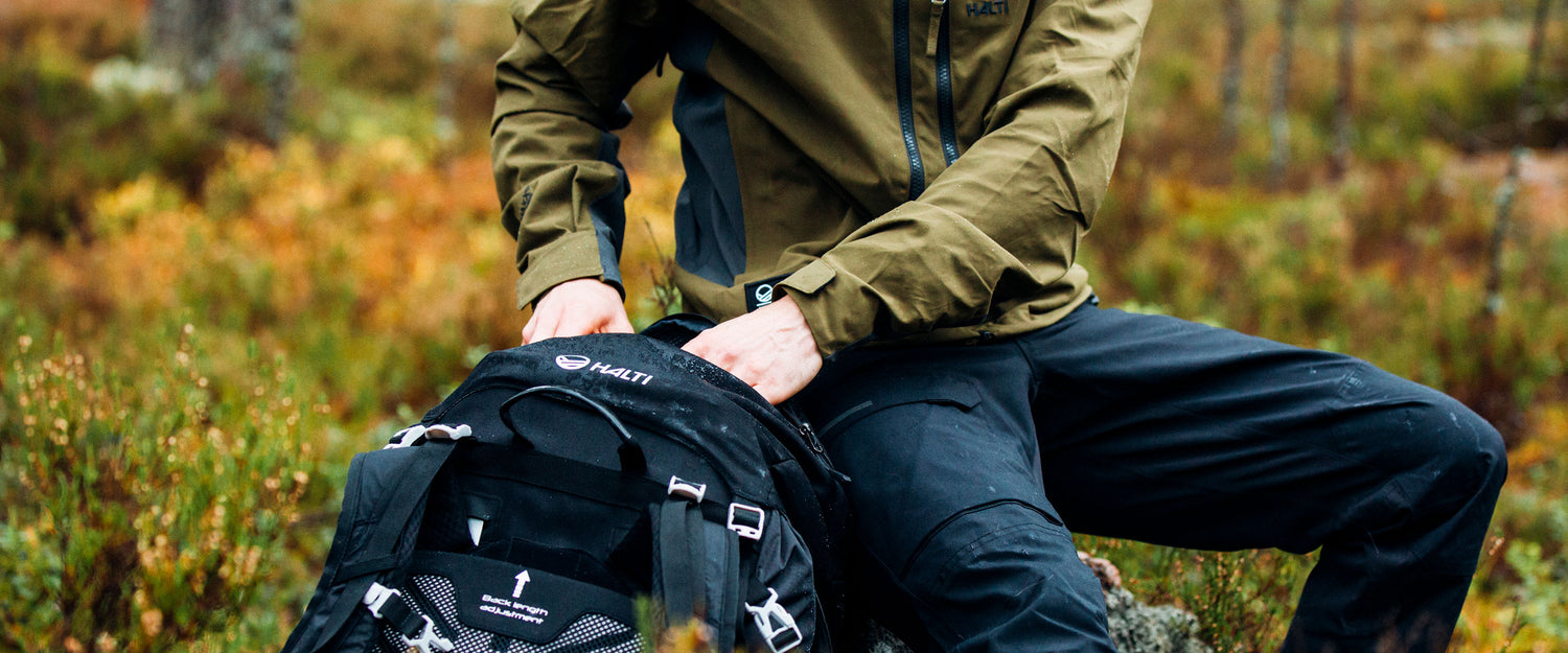 A person in an olive green jacket and black pants sits outdoors on a rock, opening a black Halti backpack surrounded by green and orange foliage.