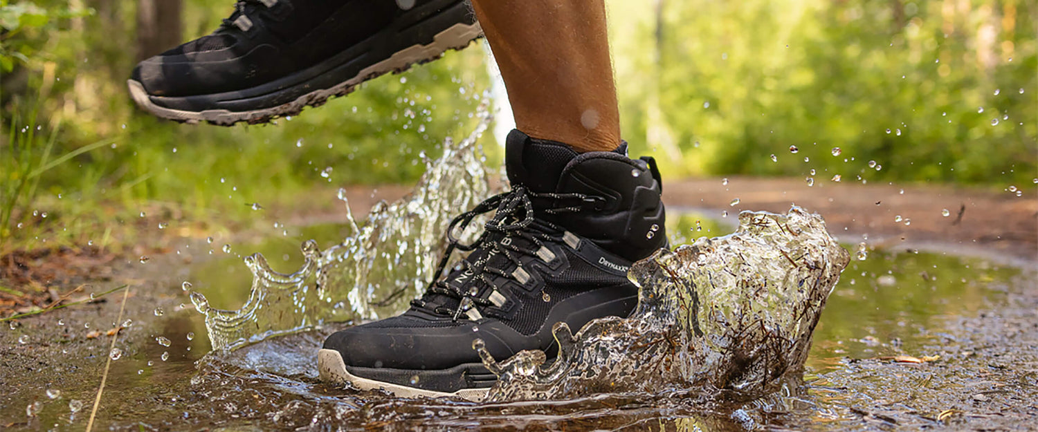 A person wearing black hiking boots steps into a shallow puddle on a forest path, causing water to splash up around their feet on a sunny day.