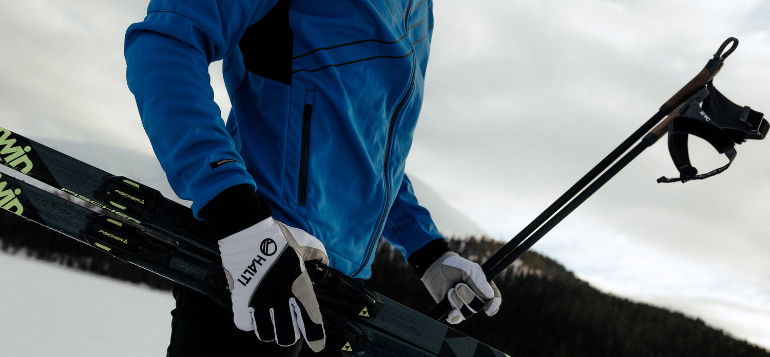 A person wearing a blue jacket and white gloves carries a pair of skis and ski poles outdoors, with a snowy landscape and trees in the background.