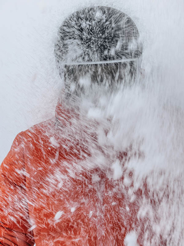A person wearing a helmet and red jacket is hit in the face with a large spray of snow, partially obscuring their face and upper body.