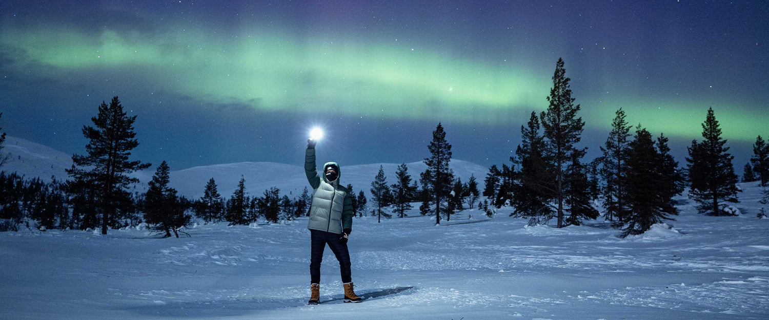 A person in a winter jacket and boots stands on snow, holding up a flashlight, with pine trees and green aurora borealis visible in the night sky above.