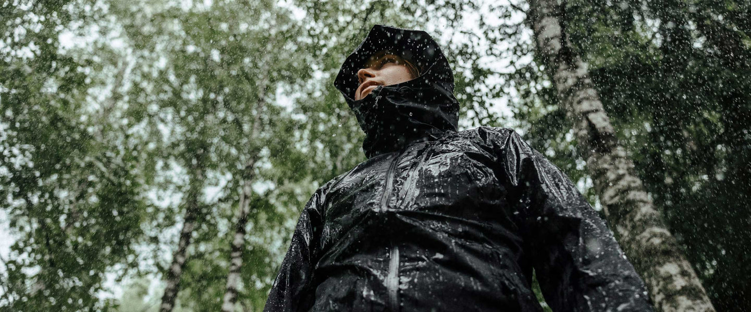 A person wearing a black rain jacket with a hood stands outdoors in a forest as rain falls, surrounded by green trees and wet foliage.