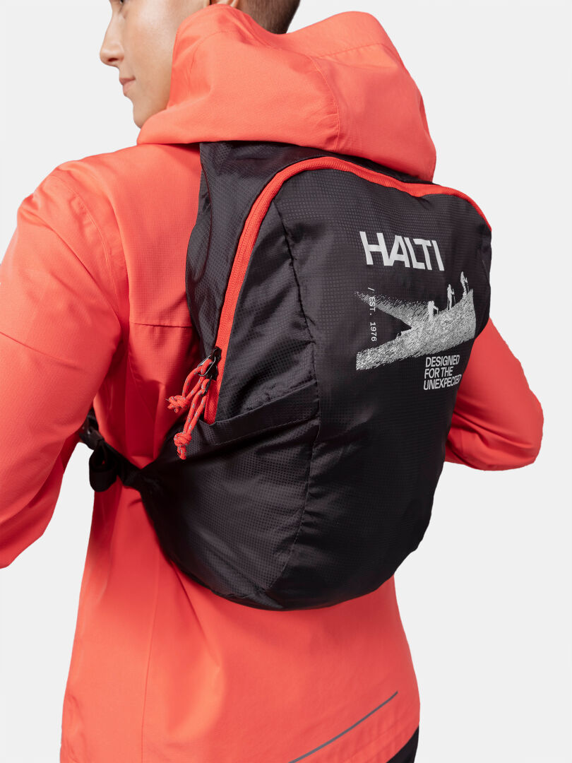 A person in an orange jacket wears a black Halti Streetpack Backpack with white text and hiker graphics on the back.