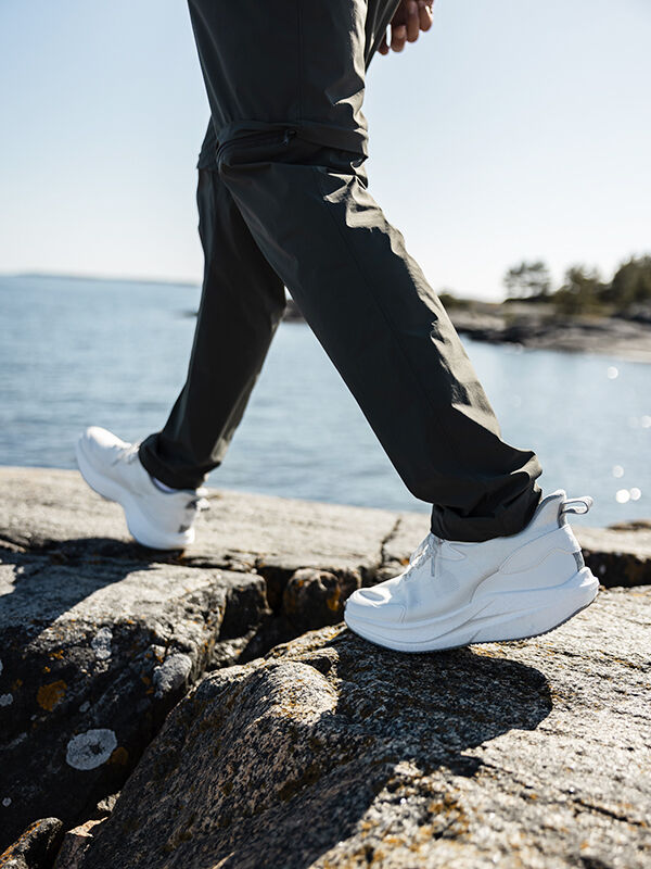 Wearing Halti's Syke Sneaker Men's and dark pants, a man walks on rocks by the water on a clear day.