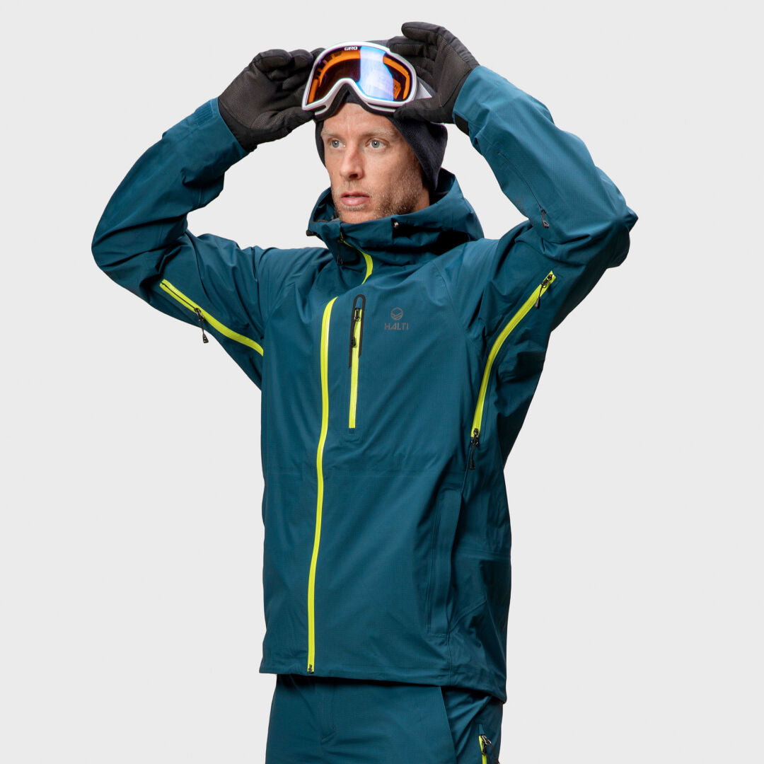 Wearing a Halti Alpine Unisex 3L DrymaxX Shell Jacket, a person puts on ski goggles against a plain light background.