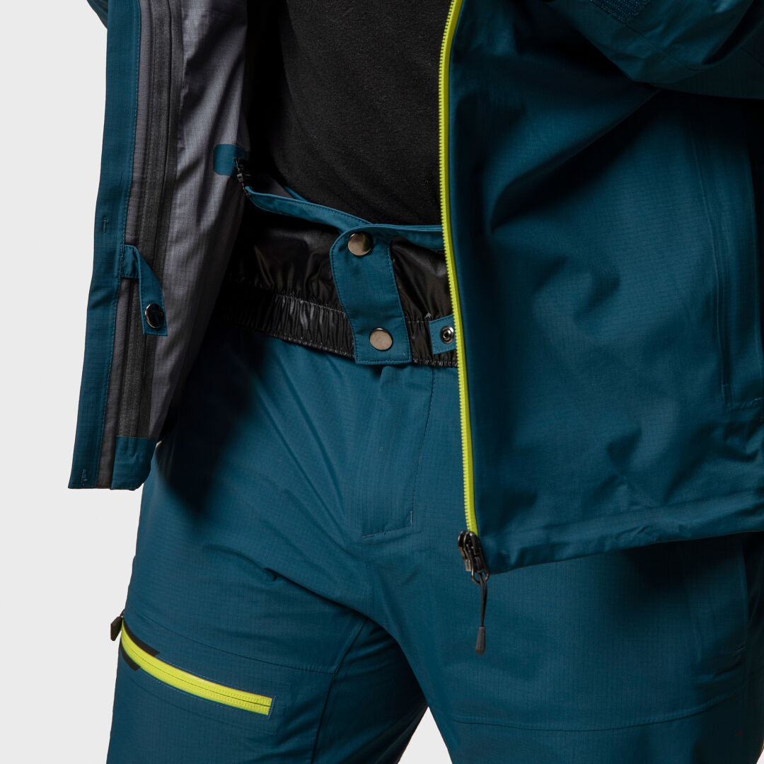 Person wearing a partially unzipped Halti Alpine Unisex 3L DrymaxX Shell Jacket and teal outdoor pants with yellow zippers.