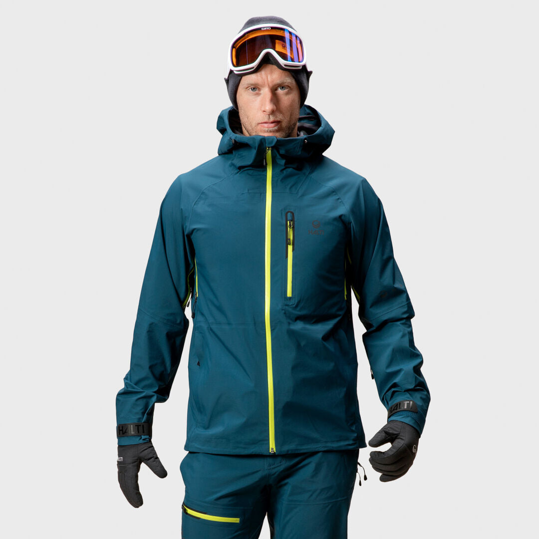 A man wears a Halti Alpine Unisex 3L DrymaxX Shell Jacket with yellow zippers, black gloves, and ski goggles, facing forward.