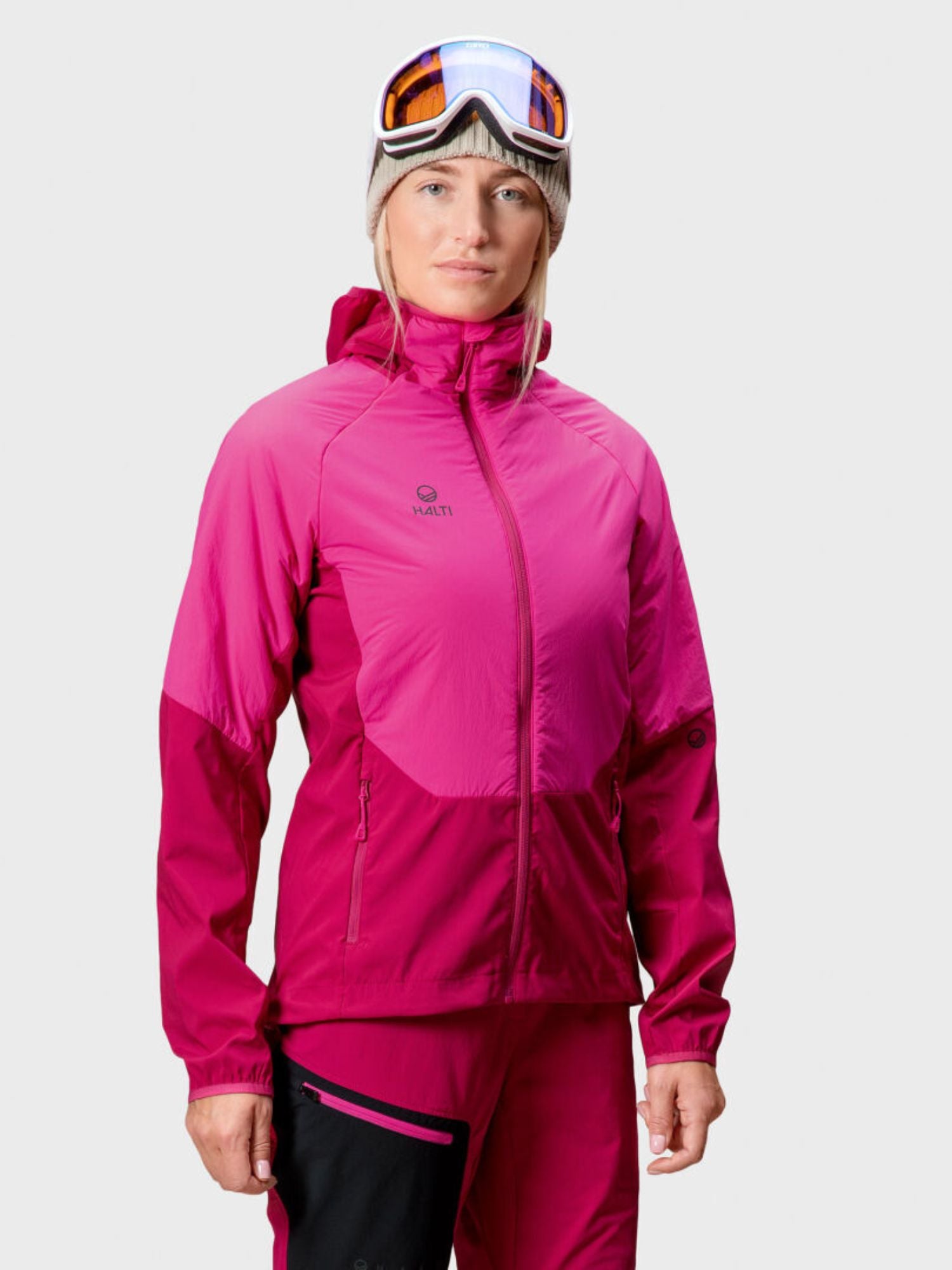 A woman wears the Halti Crust Women's Layer Jacket with matching pants, ski goggles, and a beige beanie, facing forward.