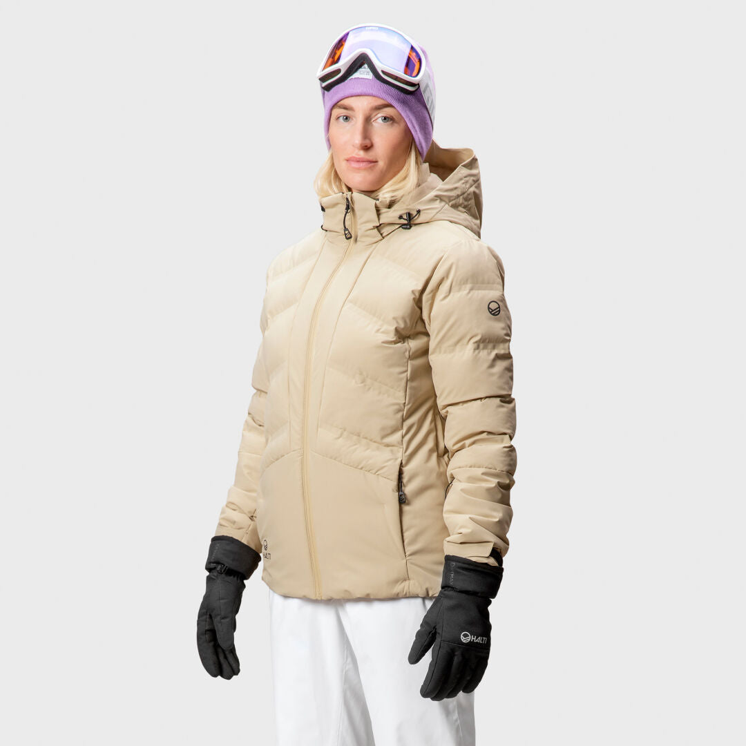 Person wearing a Halti Nordic Arcty Ski Jacket Womens in beige, black gloves, white pants, and a purple hat with ski goggles.