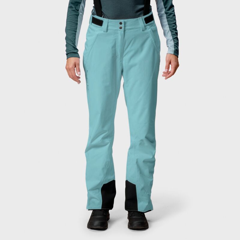 A woman in Halti Trusty DrymaxX Ski Pants in light blue with black ankle patches and a teal long-sleeve top, front view.