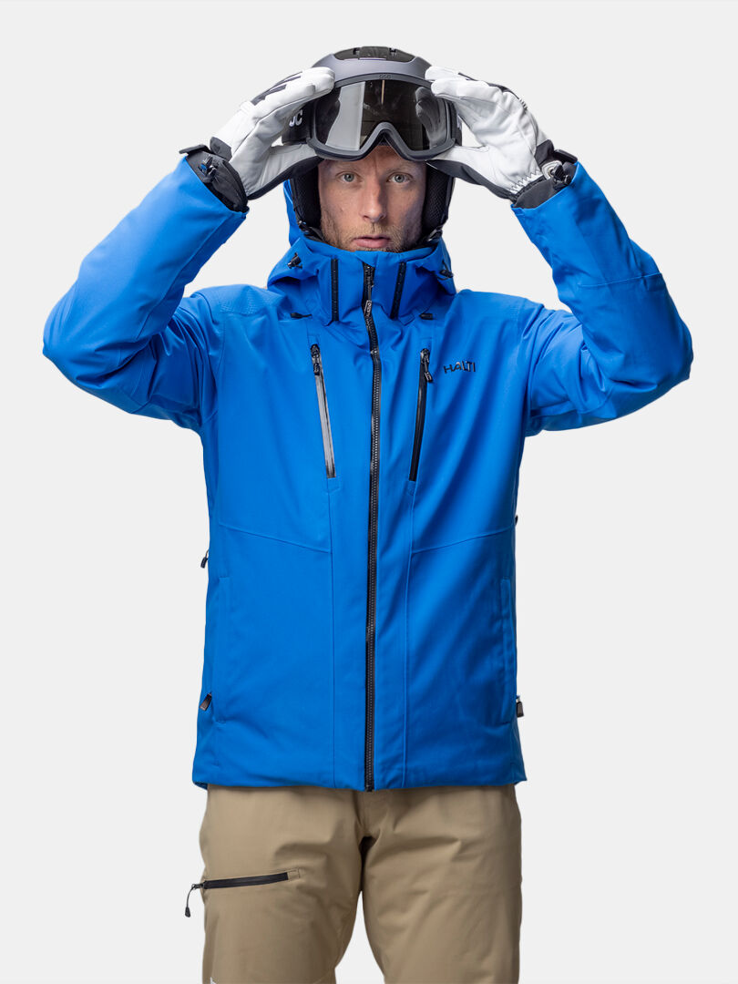 A person in a blue Halti Vertica DX Ski Jacket and tan pants adjusts ski goggles, facing forward.