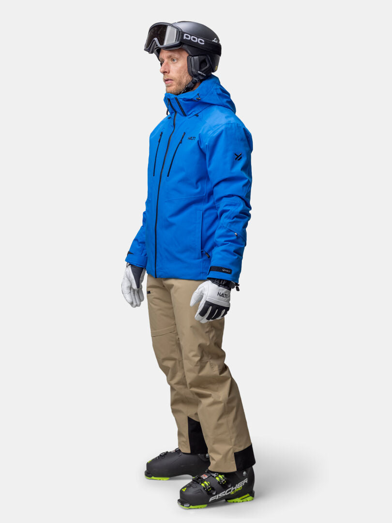 A man in a Halti Vertica DX Men's blue ski jacket, tan pants, gloves, boots, helmet, and goggles stands facing right.