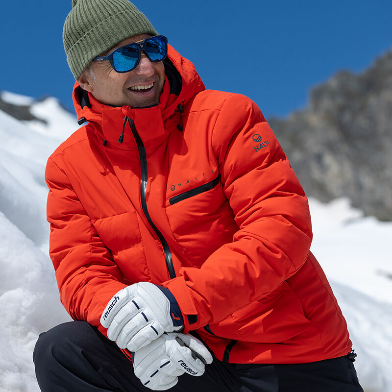 A man in a Halti Nordic Arcty Ski Jacket Mens smiles on a snowy mountain slope under a clear blue sky.