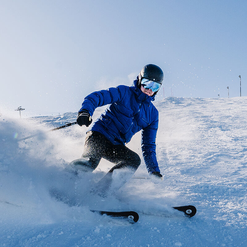 A skier in a Halti Nordic Arcty Ski Jacket Mens and helmet turns on a snowy slope beneath a clear blue sky.