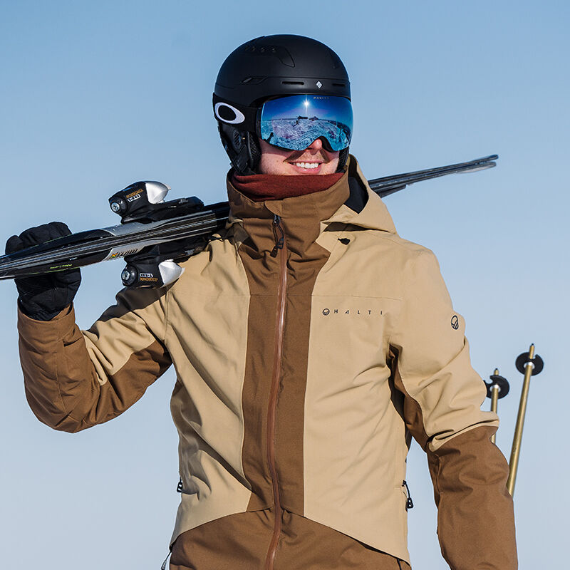 Man in Halti Planker DrymaxX Ski Jacket with helmet and goggles carries skis on shoulder under clear sky.