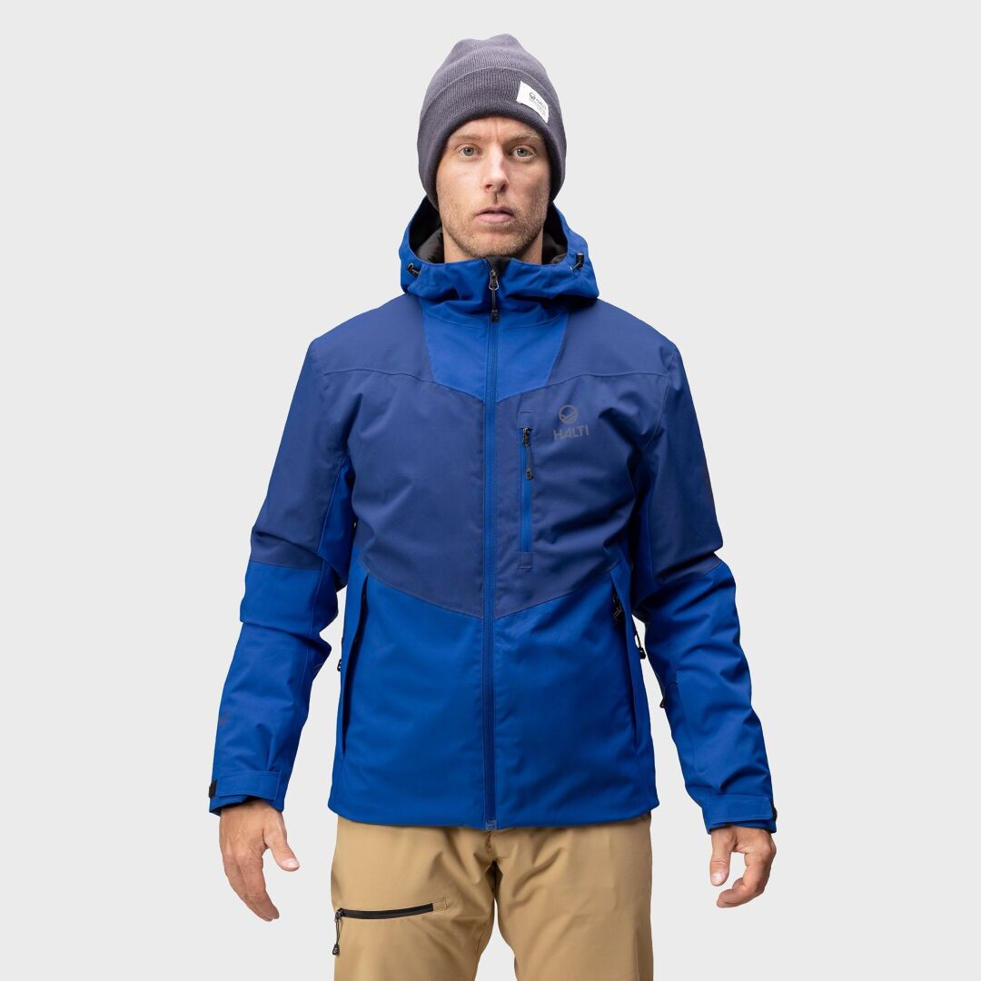 A man wears the Halti Wedeln DrymaxX Ski Jacket, tan pants, and gray beanie, standing against a plain light background.