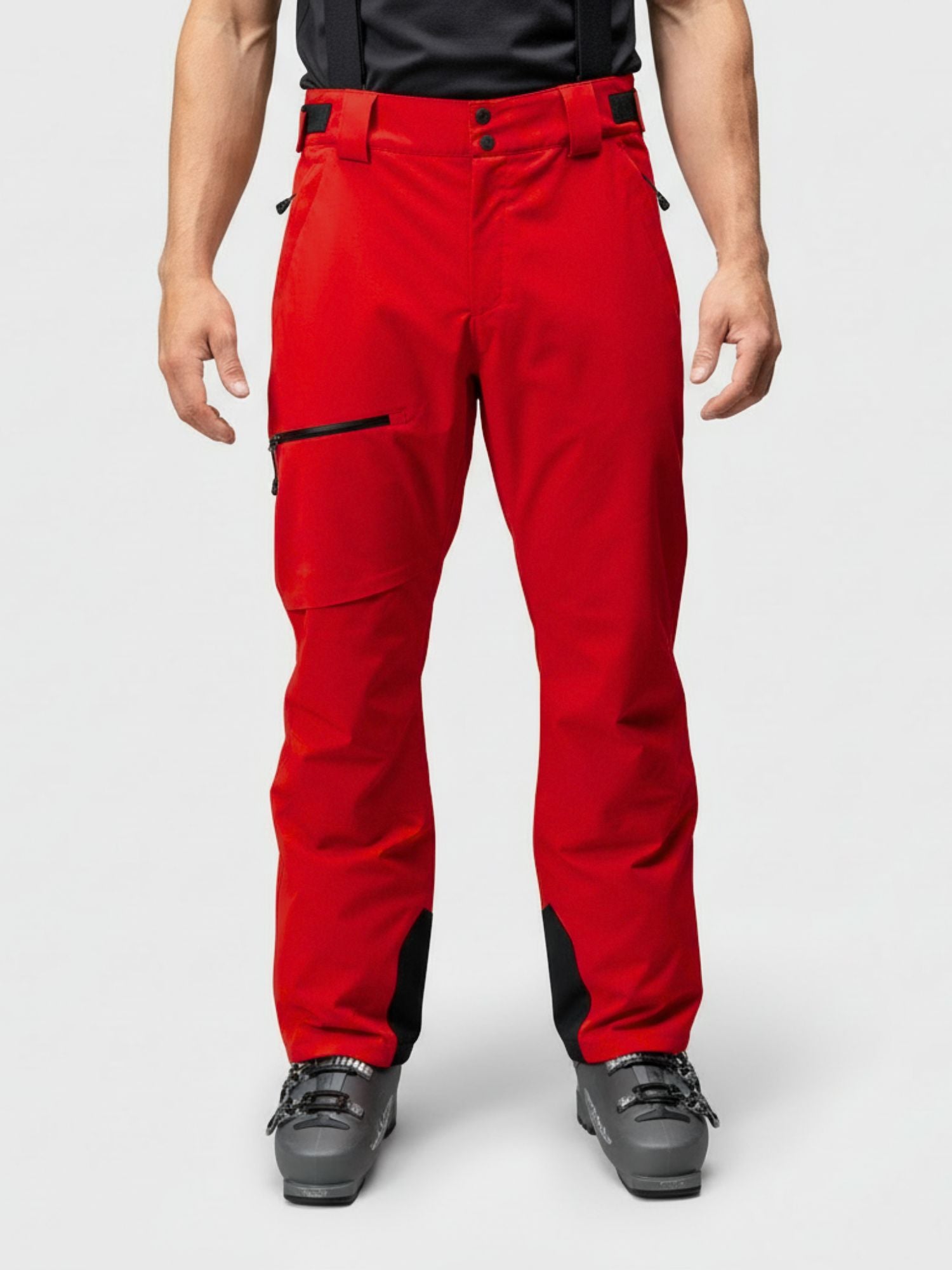Man in Halti Carvey DX bright red waterproof ski pants with black suspenders and ski boots stands against plain background.