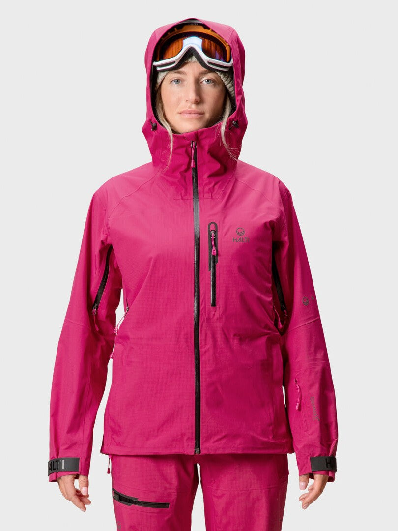 A woman wears a bright pink Halti Alpine 3L DrymaxX Shell Jacket and matching pants with ski goggles against a plain backdrop.