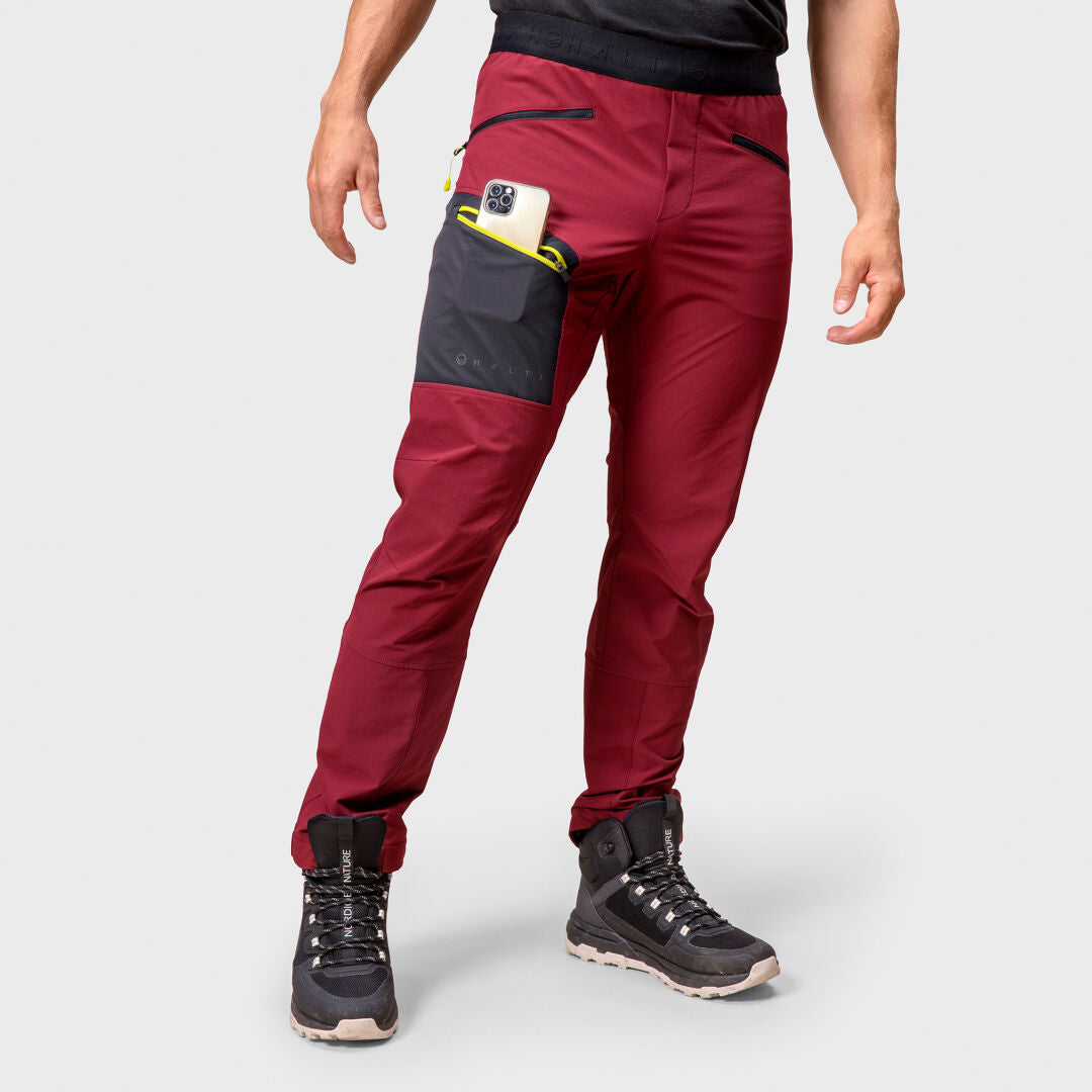 A man wears Halti Adrenaline Lite Ski Touring Pants in red and black boots, holding a phone against a white background.