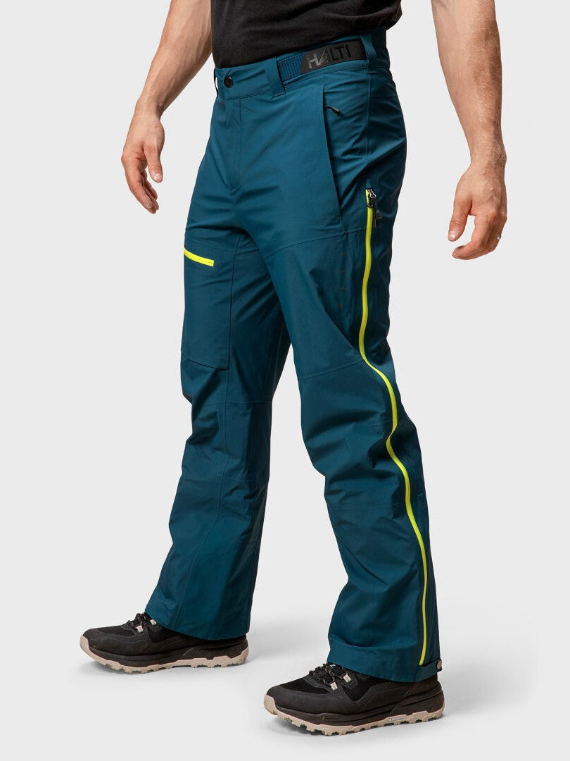 A man wears Halti Alpine 3L DrymaxX Shell Pants Men’s with yellow zippers, a black shirt, and hiking shoes.