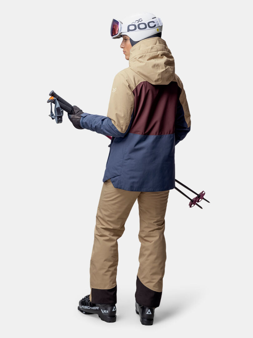 Woman in Halti Gracer DX Ski Jacket and helmet, holding ski poles, photographed from the back on plain background.