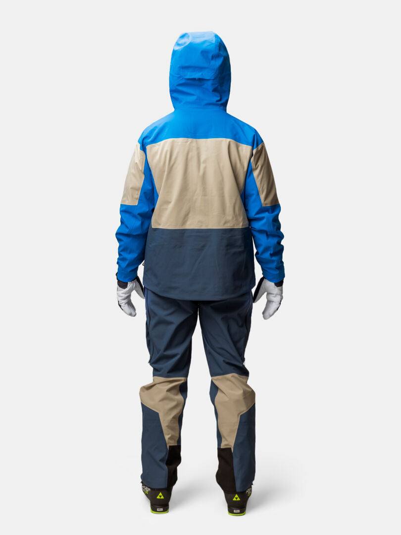 A person seen from behind wearing a blue and tan Halti Rizle Unisex 3L DX Ski Jacket, dark trousers, and white gloves.