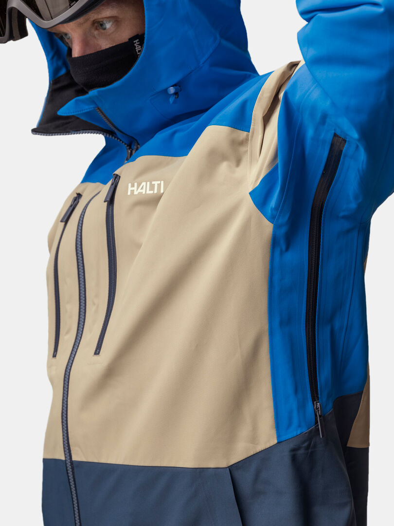 A person in a Halti Rizle 3L DX Ski Jacket zips a pocket, wearing ski goggles and a face covering.