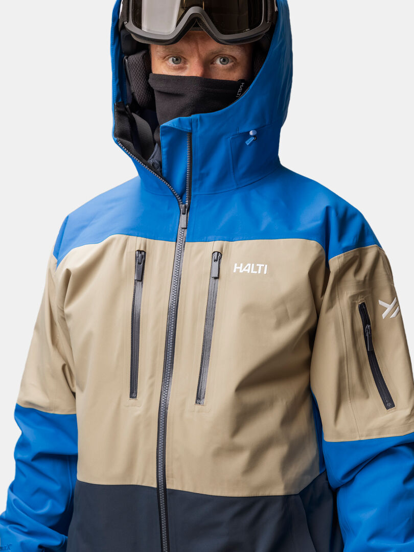 Person wearing a Halti Rizle 3L DX Ski Jacket (blue, beige, black) with hood up, ski goggles, and face covering.