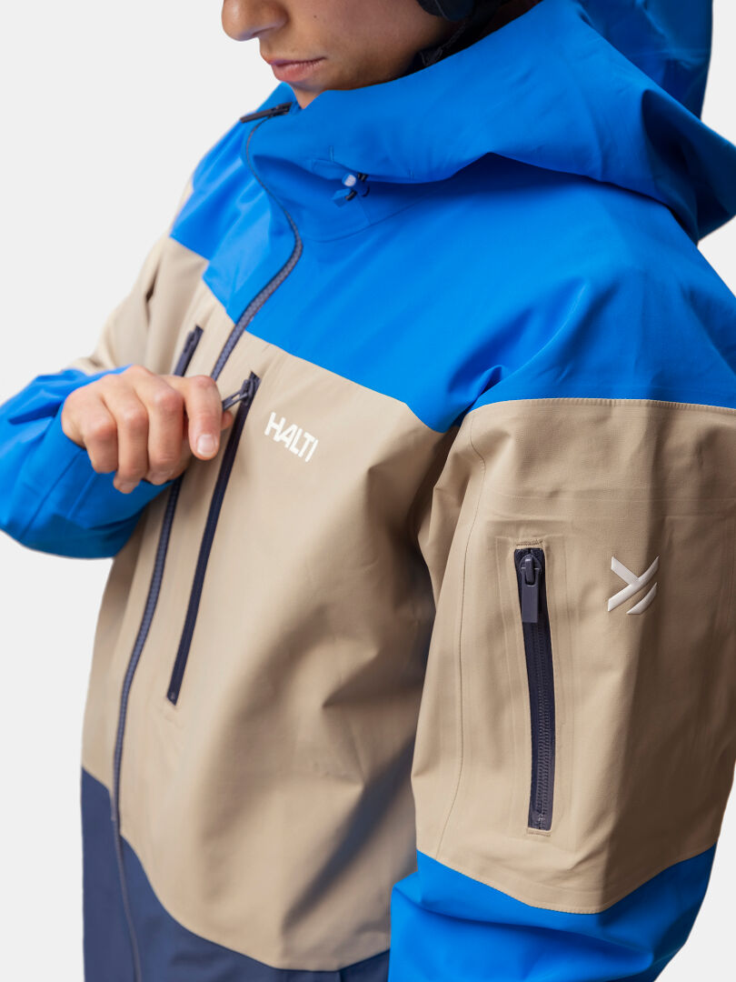 Person zipping up a Halti Rizle 3L DX Unisex ski jacket in beige and blue, logo visible on chest and sleeve.