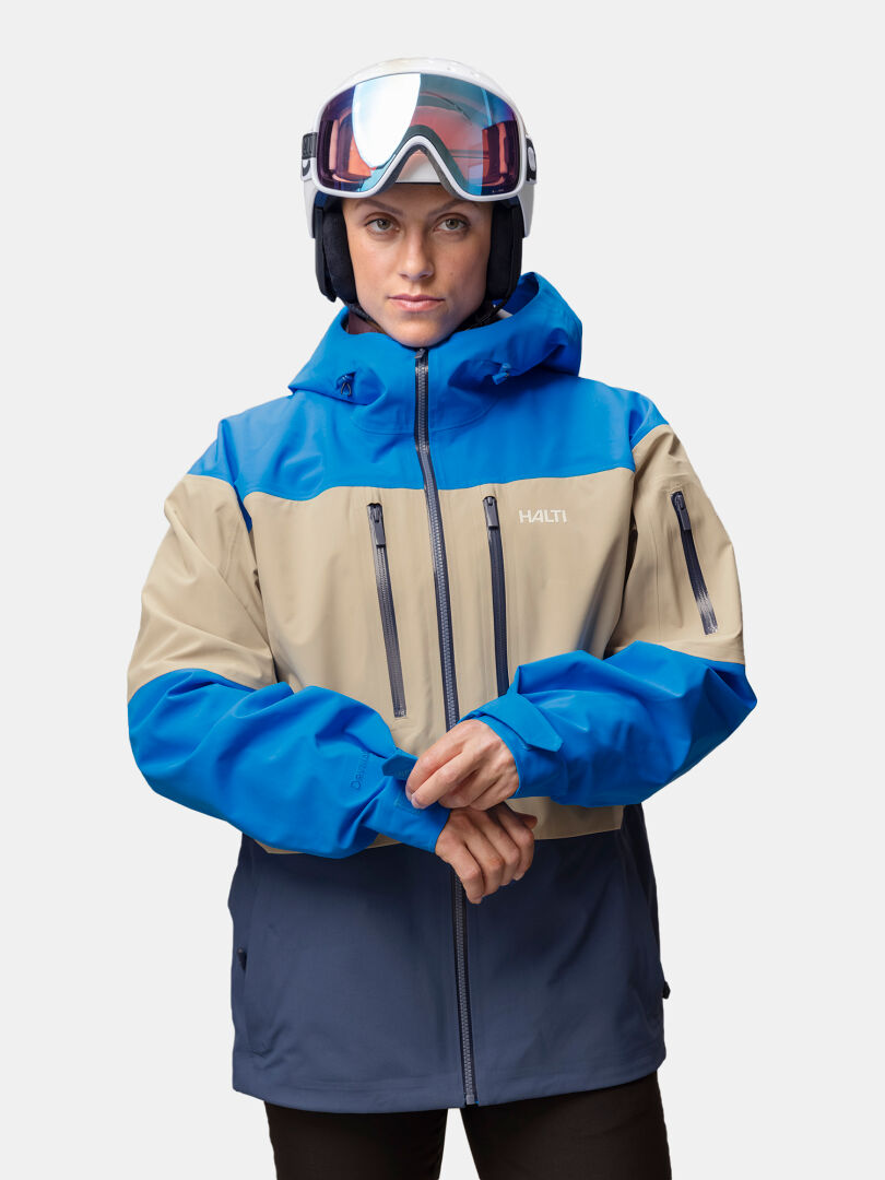 A person wearing a ski helmet and a blue and tan Halti Rizle Unisex 3L DX Ski Jacket with zippered chest pockets.