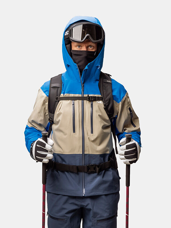 A skier wearing a Halti Rizle Unisex 3L DX Ski Jacket, helmet, goggles, face mask, and gloves while holding ski poles.