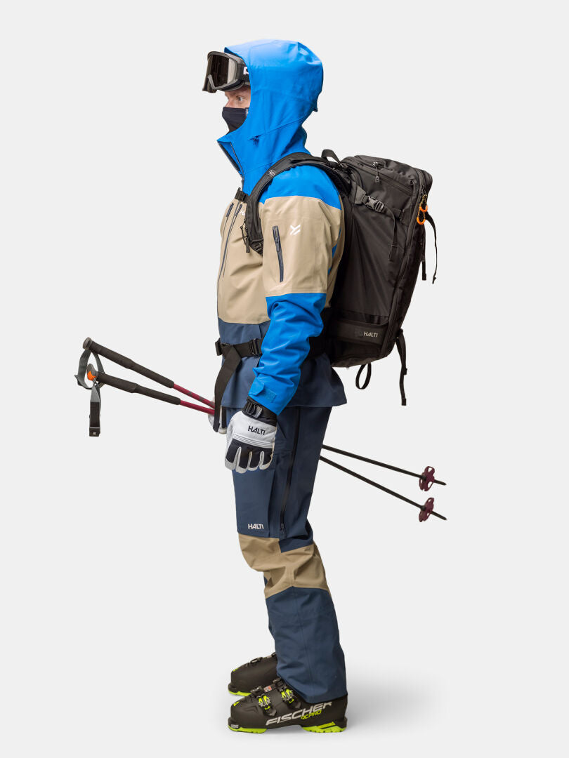 Person wearing a blue and tan Halti Rizle Unisex 3L DX Ski Jacket, pants, helmet, goggles, and backpack, holding ski poles.