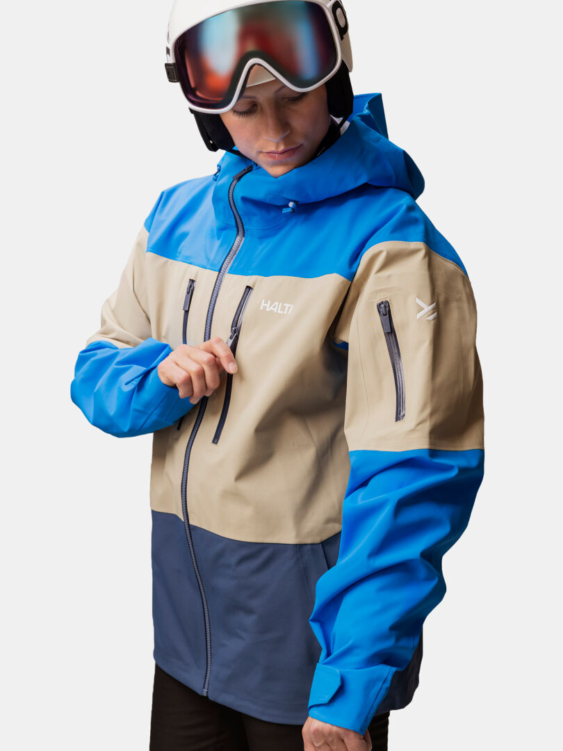 A person wearing a blue and tan Halti Rizle Unisex 3L DX Ski Jacket with a helmet and goggles.